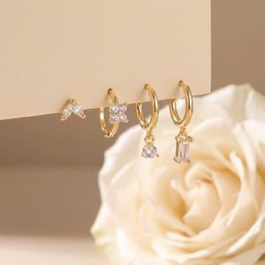 May include: A collection of gold-colored earrings with sparkling white gemstones. The earrings include small studs and hoop styles, some with dangling charms. The earrings are displayed against a neutral background with a white rose in the corner.