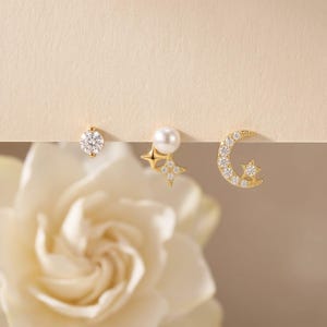 May include: A set of three gold-tone stud earrings. The first is a round clear stone, the second features a white pearl with a four-pointed star, and the third is a crescent moon shape encrusted with small clear stones and a small star accent.