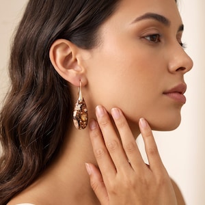 May include: A teardrop-shaped earring featuring a mottled, multi-coloured stone with shades of brown, cream, and dark amber. The stone is suspended from a simple, thin gold-toned metal hook that passes through the earlobe for secure attachment.
