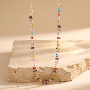 May include: A gold-tone chain necklace featuring a variety of multi-colored, multi-shaped gemstones including pink, yellow, blue, purple, and green stones. The stones are set in individual metal frames and arranged in a single, long, continuous strand.
