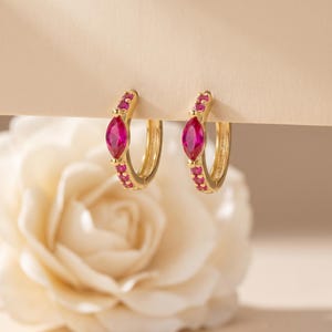 May include: A pair of gold hoop earrings featuring a central marquise-cut vibrant pink gemstone flanked by smaller round pink stones along the front of each hoop. The earrings have a polished gold finish and a hinged closure mechanism.