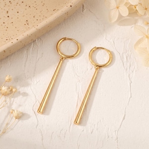May include: A pair of gold-toned hoop earrings, each featuring a smooth, cylindrical bar pendant suspended from a circular hoop. The earrings have a polished, reflective metallic finish and a simple, clean design with a hinged closure mechanism.