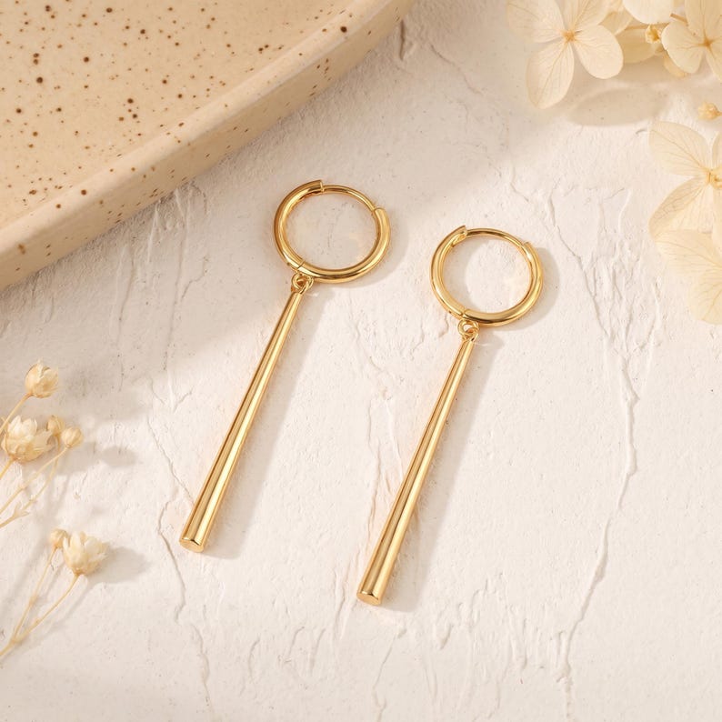May include: A pair of gold-toned hoop earrings, each featuring a smooth, cylindrical bar pendant suspended from a circular hoop. The earrings have a polished, reflective metallic finish and a simple, clean design with a hinged closure mechanism.