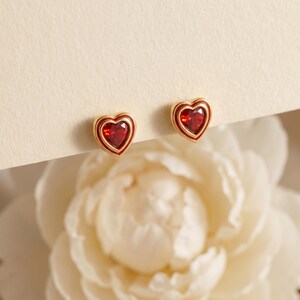 May include: A pair of gold stud earrings, each featuring a central heart-shaped red gemstone surrounded by a thin red enamel border. The earrings have a polished gold-tone metal setting that frames the vibrant red heart design clearly.