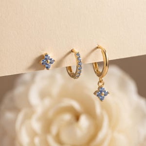 May include: A set of three gold-toned earrings featuring light blue stones. One earring is a four-petaled flower stud, one is a small hoop encrusted with light blue stones, and one is a small hoop with a dangling four-petaled flower charm.