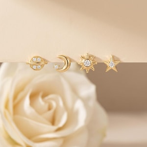 May include: Gold-toned stud earrings featuring celestial designs: a planet, crescent moon, sun, and star. Each earring has a small, sparkling gemstone. The earrings are displayed on a neutral background with a soft, blurred flower in the foreground.