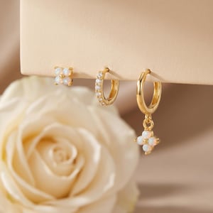 May include: A set of three gold-colored earrings. One is a stud with four white opal stones in a flower shape. The second is a small hoop with clear stones. The third is a hoop with a flower-shaped opal charm.
