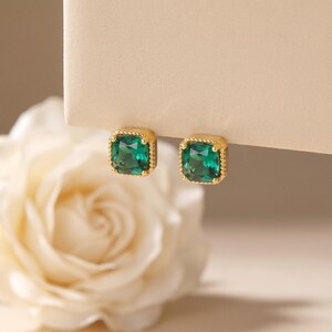 May include: A pair of gold stud earrings featuring square-cut, deep green faceted gemstones. Each stone is set within a gold frame with a delicate beaded edge, providing a textured border around the vibrant green center of the jewelry pieces.