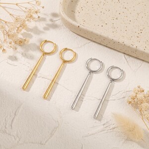 May include: Two pairs of drop earrings, one pair in a polished gold tone and one pair in a polished silver tone. Each earring consists of a circular hoop connected to a smooth, cylindrical bar pendant that hangs vertically from the hoop base.