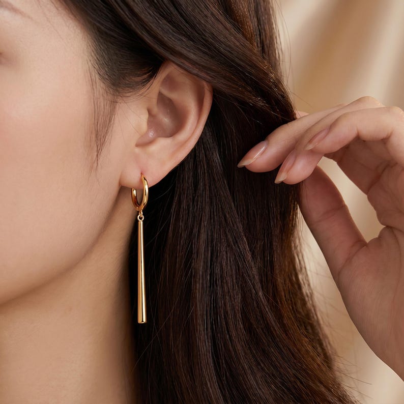 May include: A gold-toned hoop earring featuring a long, slender, cylindrical drop pendant. The earring consists of a circular hoop that connects to a smooth, tapered metal bar that hangs vertically from the base of the hoop.