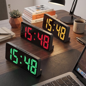 May include: Three rectangular digital alarm clocks with black frames and mirrored screens. Each clock displays the time 15:48 in bright LED digits, with one clock showing green numbers, one showing red numbers, and one showing yellow numbers.