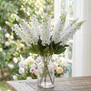 May include: A clear glass vase displays a bouquet of white flowers with green stems and leaves. The flowers have a tall, slender form and are arranged in a natural style. The vase is on a wooden table, with a blurred background of greenery and sunlight.