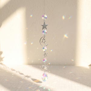 May include: A hanging suncatcher ornament featuring a vertical chain of silver-toned metal charms including stars, a crescent moon with a star cutout, and a sun, interspersed with multifaceted iridescent glass beads and a teardrop crystal.