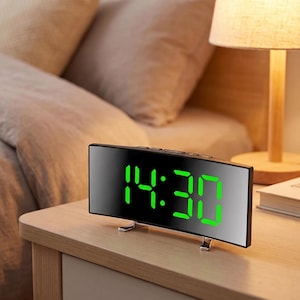 May include: A curved digital alarm clock with a reflective black surface and a bright green LED display showing the time as 14:30. The clock features a sleek, rectangular design with small metallic support feet on the bottom edge for stability.