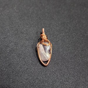 以下が含まれることがあります： 銅線で包まれた、磨かれた薄紫色の石を使った手作りのペンダント。ワイヤーは石の周りに複雑に織り込まれ、ユニークで芸術的なデザインを作り出しています。ペンダントは、同じ銅線の小さなループから吊り下げられています。