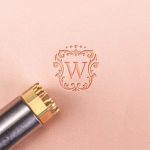 Könnte beinhalten: Ein Lederwerkzeug mit Messingkopf und silbernem Griff befindet sich in der Nähe eines geprägten Buchstabens "W" in einem verzierten Wappen. Das Wappen ist von dekorativen Verzierungen und Sternen umgeben, alles in einem warmen, rötlich-braunen Farbton.