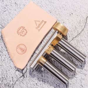 May include: A set of leather working tools and a light tan leather piece. The tools have silver and gold metal components. The leather piece has the text "Saphirex", "W", and "ANNIC NOBLE" with an anchor.