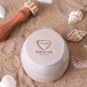 May include: A beige ceramic bowl with the name "MICHELLE" and a bowl symbol stamped on the bottom. The bowl is sitting on sand, with seashells and a wooden and brass tool in the background.