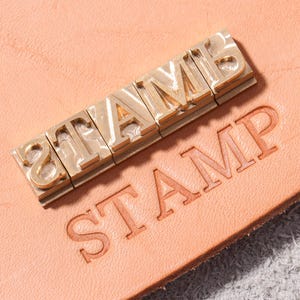 May include: A set of five individual brass metal letter stamps spelling the word STAMP, positioned above a light tan leather surface that features the word STAMP clearly embossed into the material in a matching serif font style.