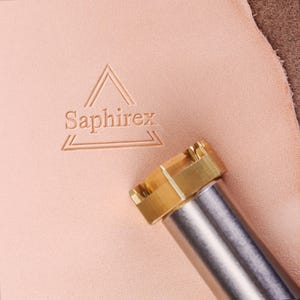 Puede incluir: Primer plano de una herramienta de estampado de cuero con cabezal de latón y mango plateado. La herramienta está presionada sobre un trozo de cuero marrón claro, creando un diseño en relieve con la palabra "Saphirex" dentro de un triángulo.