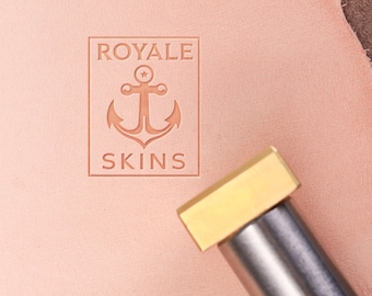 Może przedstawiać: Zbliżenie stempla skórzanego z napisem "ROYALE SKINS" i wzorem kotwicy. Stempel jest wciśnięty w kawałek jasnoróżowej skóry. Metalowe narzędzie z mosiężną główką znajduje się na pierwszym planie.