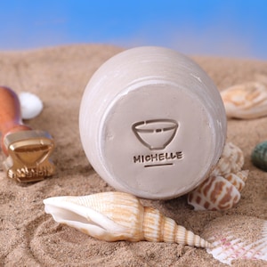 May include: A beige ceramic bowl with a stamped bowl design and the name "MICHELLE" on the base. The bowl is on sand, with seashells and a wooden stamp in the background. The sky is blue.