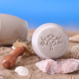 May include: A light beige ceramic stamp with a wooden handle and a tree design, alongside a matching ceramic jar. The items are on a sandy surface with seashells and a pink rock, against a blue background.