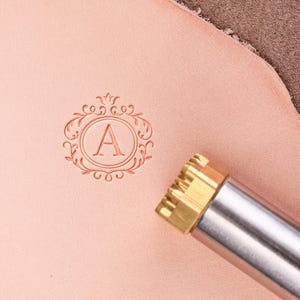 Puede incluir: Una herramienta de estampado de cuero con cabezal de latón y mango plateado. La herramienta ha estampado la letra "A" dentro de un diseño circular ornamentado sobre cuero rosa. El diseño incluye elementos florales y de volutas.