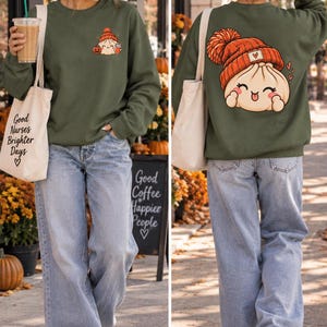 May include: An olive green crewneck sweatshirt featuring a small graphic on the front left chest of a dumpling wearing an orange beanie and holding a pumpkin. The back displays a large version of the same dumpling character with a heart icon.
