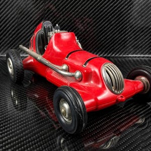 May include: A vintage-style red toy race car featuring a prominent silver front grille, black rubber tires with silver hubs, and an exposed engine detail behind the driver seat. The body is bright red with black accents and a metallic exhaust pipe.