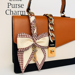 May include: A brown, black, and white purse with a gold chain and a decorative bow purse charm. The charm has a cream-colored bow with a brown houndstooth pattern. The purse has a gold lock with the brand name "ALDO". The text "Chic Purse Charm" is visible.