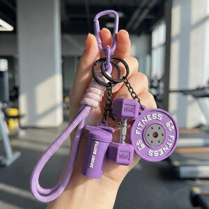 May include: A purple keychain featuring a carabiner, a braided cord loop, and three charms: a shaker cup labeled SPORT BOTTLE, a dumbbell marked 10KG, and a circular weight plate inscribed with FITNESS and 45 LB. The charms hang from black chains.