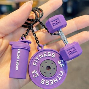 May include: A purple keychain featuring three charms: a shaker cup labeled SPORT BOTTLE, a circular weight plate marked FITNESS 45 LB, and a dumbbell with 10KG printed on each end. The charms are attached to a black metal ring by black chains.