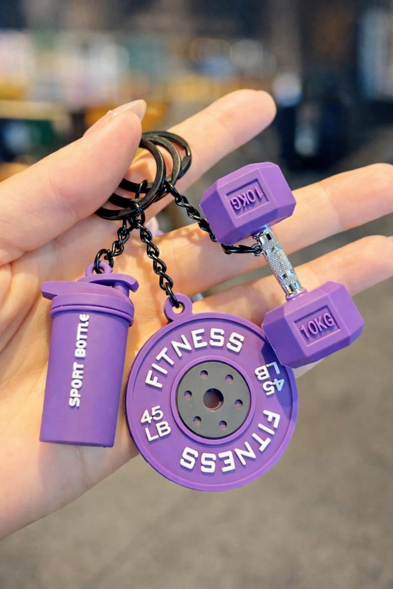 May include: A purple keychain featuring three charms: a shaker cup labeled SPORT BOTTLE, a circular weight plate marked FITNESS 45 LB, and a dumbbell with 10KG printed on each end. The charms are attached to a black metal ring by black chains.