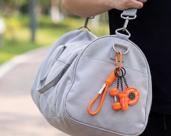 May include: A light gray duffel bag with a shoulder strap and a side pocket. Attached to the metal D-ring is a bright orange carabiner with a coiled cord, a miniature orange dumbbell, a small orange water bottle, and a round orange weight plate.