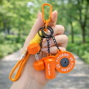 May include: An orange carabiner keychain featuring a yellow cord loop and three charms: an orange dumbbell, an orange shaker cup labeled SPORT BOTTLE, and an orange circular weight plate labeled FITNESS 45 LB with black center detailing.