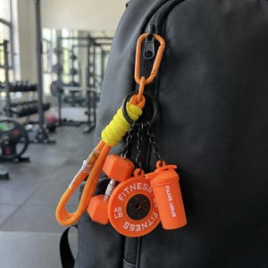 May include: A black backpack features an orange carabiner keychain with a yellow cord wrap. Attached are miniature fitness charms: an orange dumbbell, a circular orange weight plate labeled 45 LB FITNESS, and an orange sport bottle.
