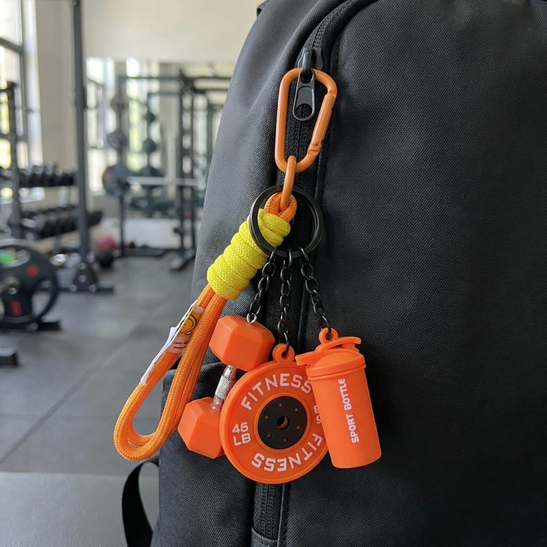 May include: A black backpack features an orange carabiner keychain with a yellow cord wrap. Attached are miniature fitness charms: an orange dumbbell, a circular orange weight plate labeled 45 LB FITNESS, and an orange sport bottle.