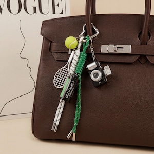May include: A dark brown leather handbag featuring a silver-toned metal keychain attachment. The keychain includes a miniature tennis racket, a neon yellow tennis ball, a black Portra 200 film canister, and a small black camera charm.