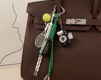 May include: A dark brown leather handbag featuring a silver-toned metal keychain attachment. The keychain includes a miniature tennis racket, a neon yellow tennis ball, a black Portra 200 film canister, and a small black camera charm.
