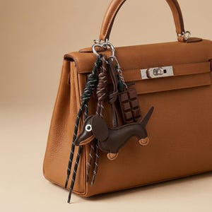 Puede incluir: Un bolso de cuero color canela con asa superior y herrajes plateados. En el asa cuelga una anilla plateada en forma de corazón con un dije de perro salchicha marrón oscuro, un dije de barra de chocolate y dos cordones trenzados.