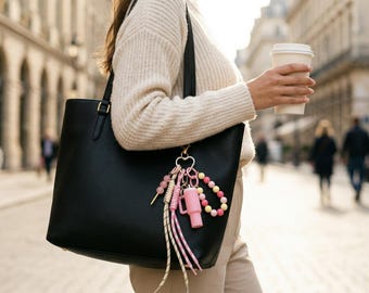 May include: A black leather tote bag featuring a heart-shaped keychain accessory. The keychain includes a miniature pink tumbler, a beaded strand with pink and yellow spheres, and two braided fabric cords in pink and white patterns.