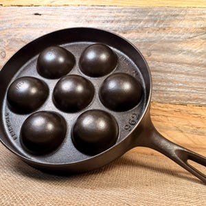 May include: A cast iron pan with seven rounded molds, ideal for making aebleskivers. The pan has a long handle with a hole for hanging. The pan is dark brown and has the number "962" stamped on it.