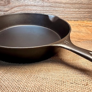 May include: A dark brown cast iron skillet with a long handle featuring an oval opening. The skillet has a wide, shallow cooking surface and a small spout for pouring. The handle is curved for easy gripping. The skillet is designed for stovetop or oven use.