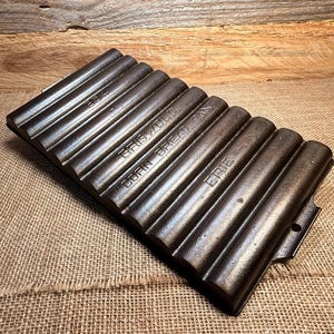 May include: A cast iron Griswold corn bread pan with a ribbed design. The pan is dark gray and has the words "Griswold", "Corn Bread Pan", and "Erie" embossed on it. The pan is on a burlap surface.