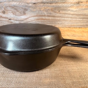 May include: A black cast iron Dutch oven with a lid. The cookware has a rounded base and a handle on each side. The lid fits snugly on top. The Dutch oven is designed for cooking.