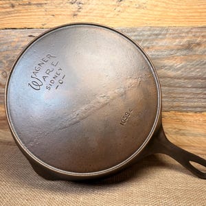 May include: A vintage cast iron Wagner Ware Sidney -0- skillet, viewed from the bottom. The dark metal pan features a handle with a hole for hanging. The text "Wagner Ware Sidney -0-" and "1059 c" are visible.