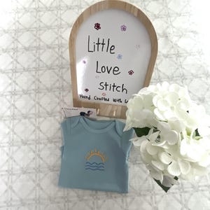 Può includere: Un body per neonati azzurro con un sole giallo ricamato che sorge sopra linee ondulate blu. Dietro il body si trova un cartello in legno ad arco con il testo Little Love Stitch Hand Crafted with Love e un bouquet di ortensie bianche.