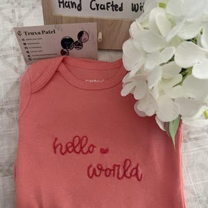 Può includere: Un body per neonati color corallo con il testo ricamato "hello world" in filo rosso. Il capo presenta uno scollo rotondo e maniche corte. Un piccolo cuore rosso è cucito tra le due parole sulla parte anteriore del tessuto.