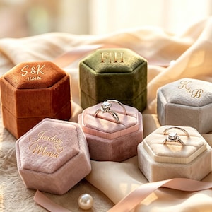 May include: A collection of six hexagonal velvet ring boxes in rust, olive green, light gray, and dusty rose colors. The boxes feature gold-embossed text including S&K 11.24.26, F|H, K&B, and Jordan Maria with a heart. Two silver rings are displayed on the boxes.